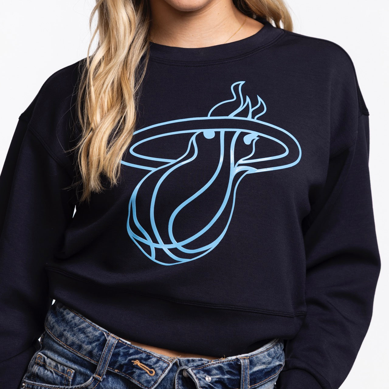 A person models the Concepts Sports Miami HEAT Original Vice Nights Cropped Women's Crewneck, featuring a light blue logo on black. The cropped fit and polyester-modal-spandex blend offer style and comfort. Worn with blue jeans; face is partially out of frame.