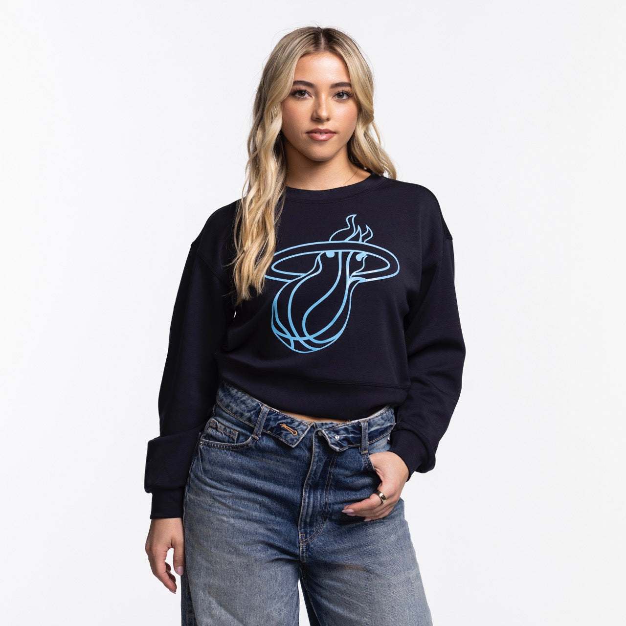 A woman with long blonde hair models the Concepts Sports Miami HEAT Original Vice Nights Cropped Women's Crewneck, a black cropped sweatshirt with a blue logo, paired with high-waisted blue jeans against a plain white background.