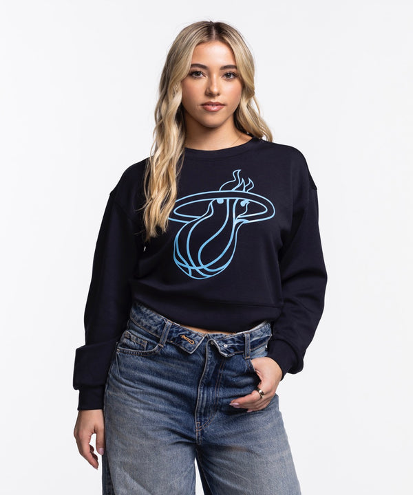 A woman with long blonde hair models the Concepts Sports Miami HEAT Original Vice Nights Cropped Women's Crewneck, a black cropped sweatshirt with a blue logo, paired with high-waisted blue jeans against a plain white background.