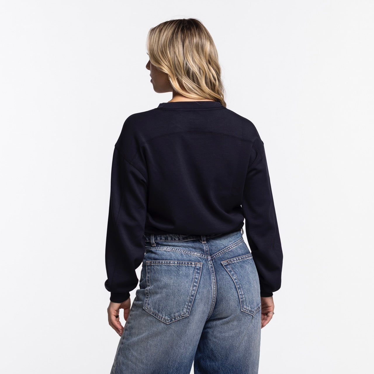 A person with wavy blonde hair stands with their back to the camera, wearing a Concepts Sports Miami HEAT Original Vice Nights Cropped Women's Crewneck and blue jeans against a plain white background.