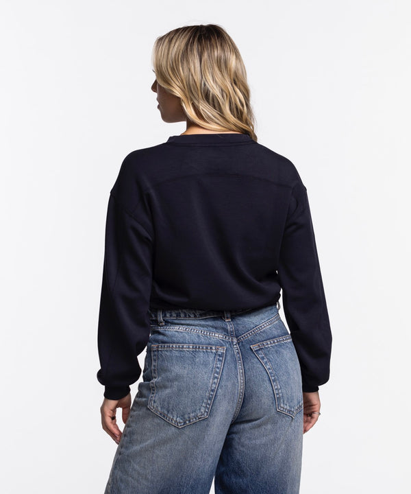 A woman with long blonde hair models the Concepts Sports Miami HEAT Original Vice Nights Cropped Women's Crewneck, a black cropped sweatshirt with a blue logo, paired with high-waisted blue jeans against a plain white background.