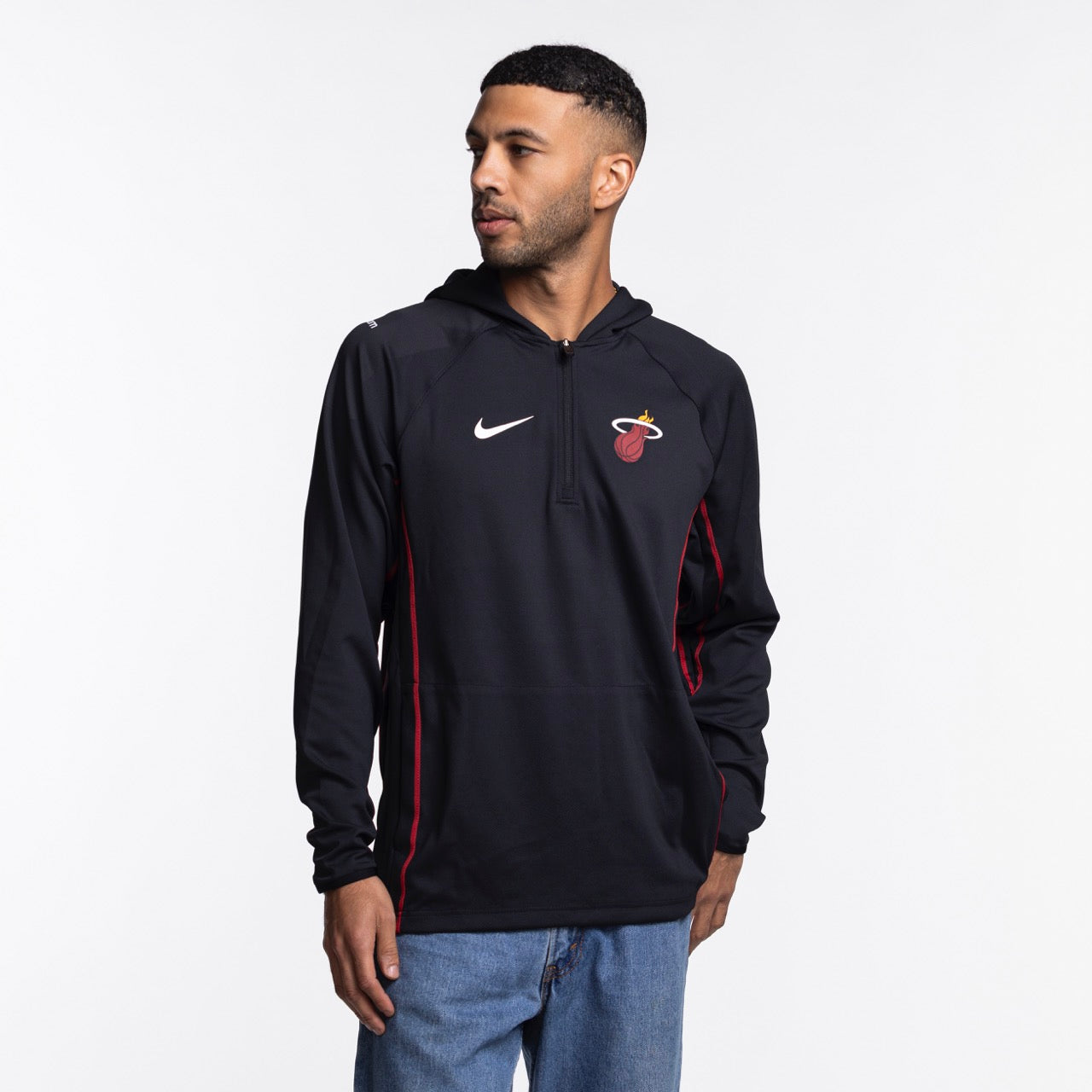 A man wears the Nike Miami HEAT On-Court 1/4 Zip Hoodie, featuring a black design with team and Nike logos, standing against a white background and looking left with one hand in his pocket.