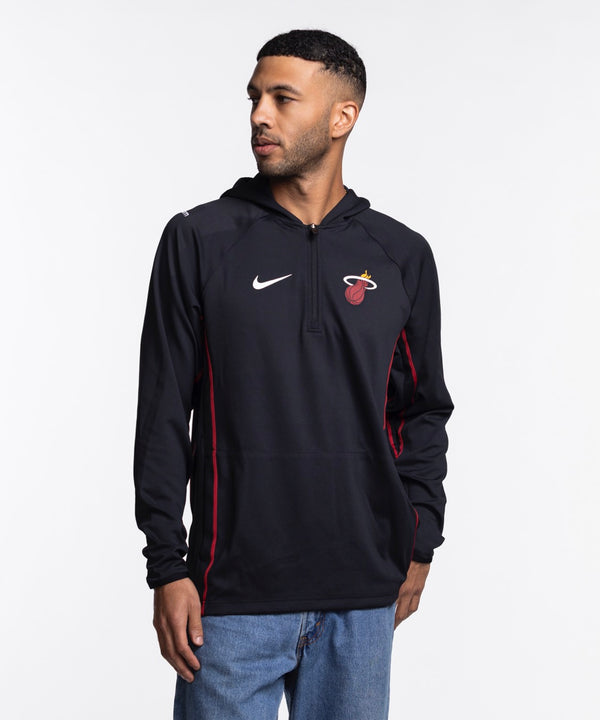 A man wears the Nike Miami HEAT On-Court 1/4 Zip Hoodie, featuring a black design with team and Nike logos, standing against a white background and looking left with one hand in his pocket.