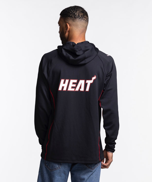 A man wears the Nike Miami HEAT On-Court 1/4 Zip Hoodie, featuring a black design with team and Nike logos, standing against a white background and looking left with one hand in his pocket.