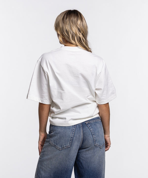 A woman with long blonde hair wears the Nike Miami HEAT Original Vice Nights Women's Tee, a loose fit white cotton shirt with blue and pink lettering by Nike, paired with blue jeans, against a plain white background. Wash with like colors recommended.