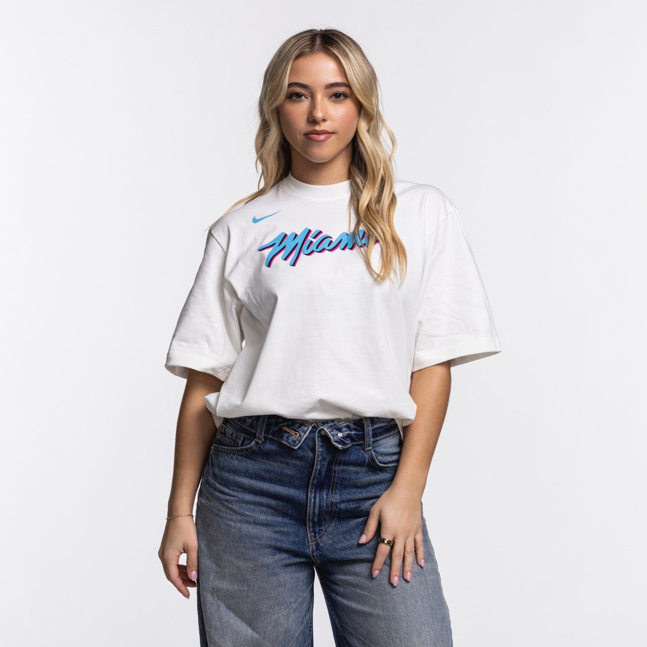 A woman with long blonde hair wears the Nike Miami HEAT Original Vice Nights Women's Tee, a loose fit white cotton shirt with blue and pink lettering by Nike, paired with blue jeans, against a plain white background. Wash with like colors recommended.