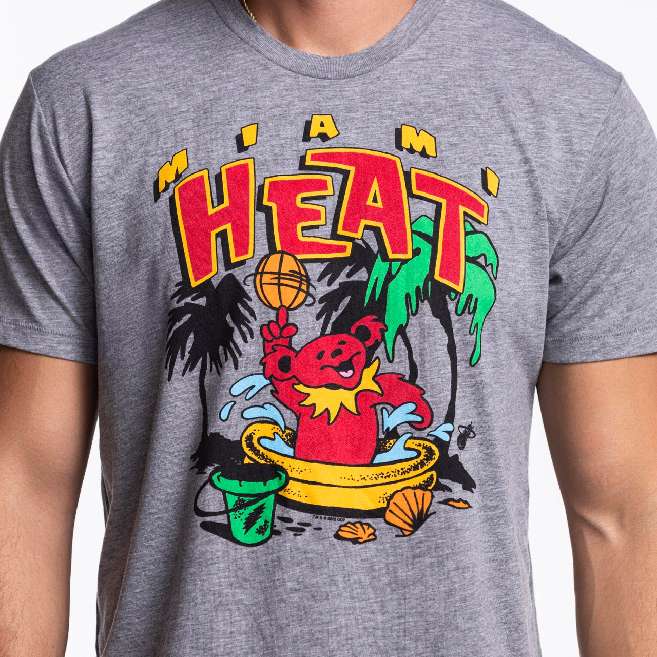 A person wears the Homage Miami HEAT Grateful Dead Bear Tee by Homage, a gray standard fit shirt with a colorful Heat logo and cartoon bear by a pool. Made from soft polyester cotton rayon, this tee is machine wash cold.