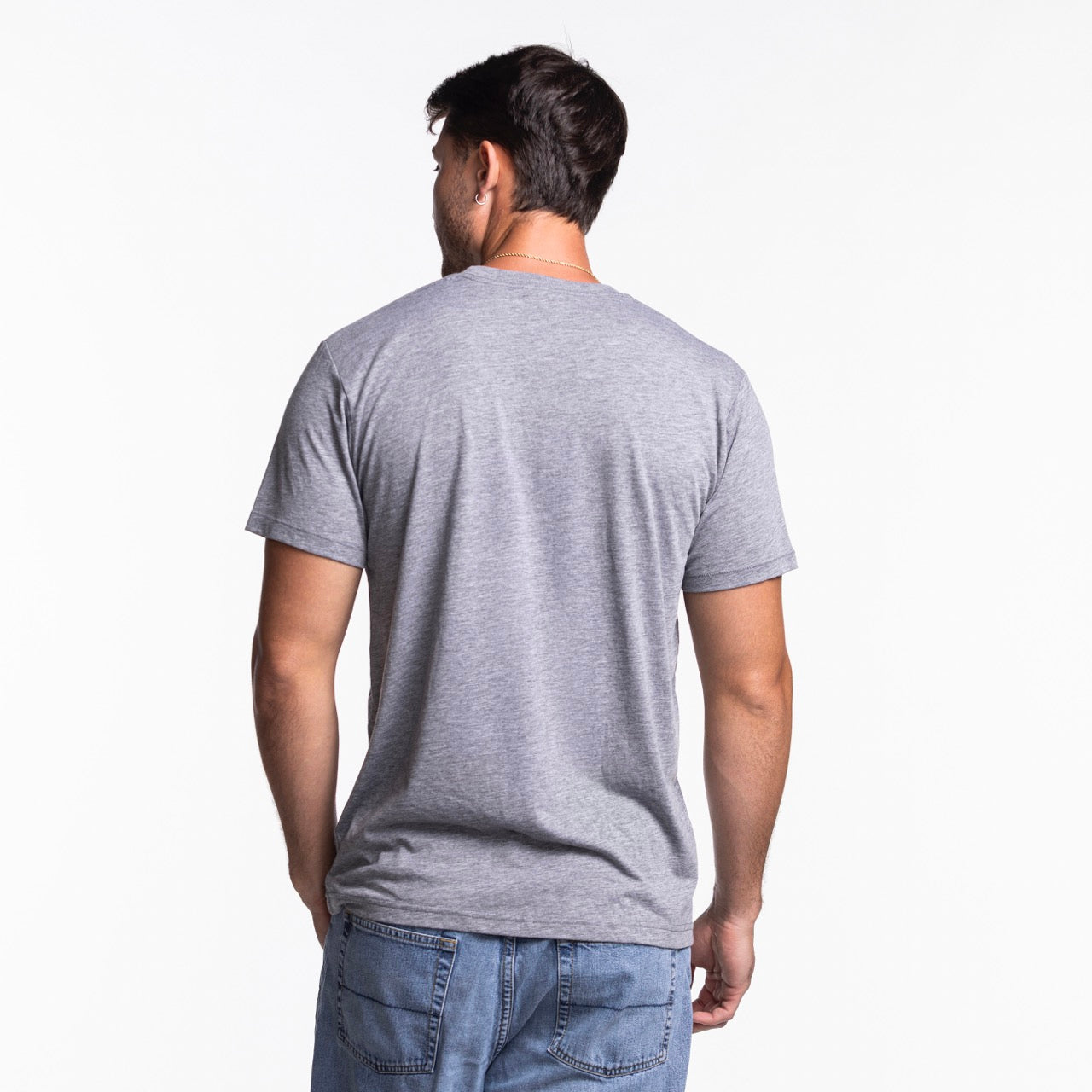A man with short dark hair faces away from the camera, wearing a Homage Miami HEAT Grateful Dead Bear Tee by Homage in gray, styled with light blue jeans against a white background. The shirt features a soft polyester cotton rayon blend.