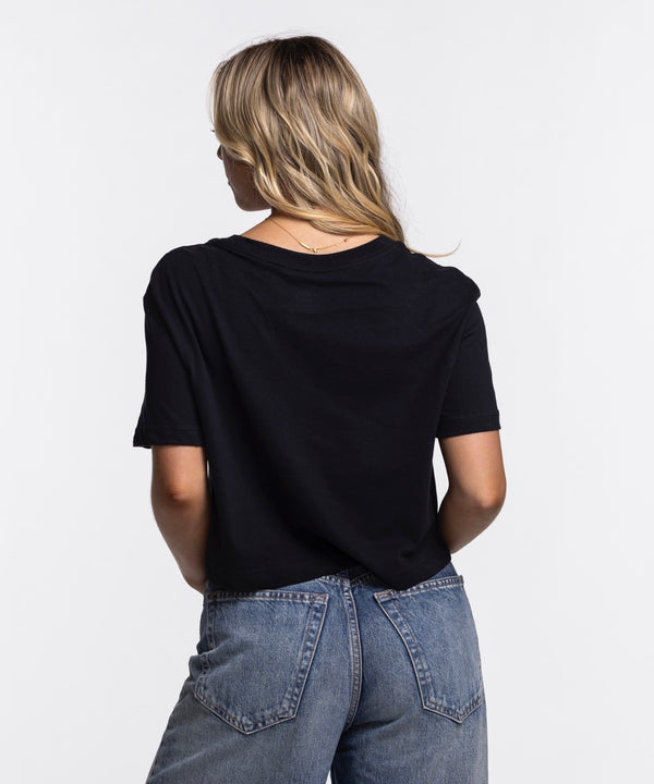 A young woman with long blonde hair smiles while wearing the Nike Miami HEAT Original Vice Nights Cropped Women's Tee, a black, soft cotton-polyester shirt with colorful “Miami” lettering, paired with blue jeans against a plain white background.