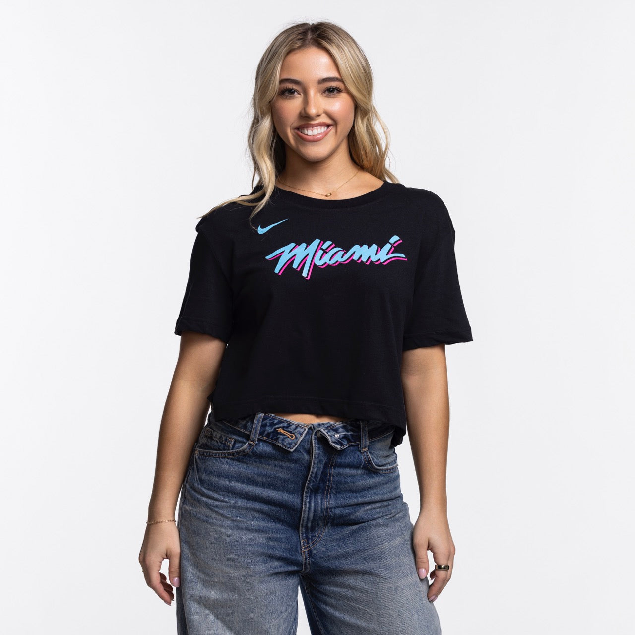 A young woman with long blonde hair smiles while wearing the Nike Miami HEAT Original Vice Nights Cropped Women's Tee, a black, soft cotton-polyester shirt with colorful “Miami” lettering, paired with blue jeans against a plain white background.