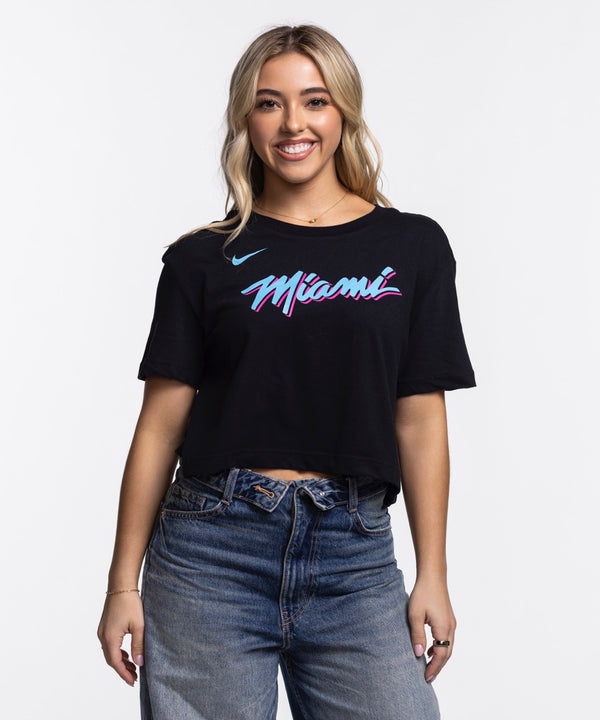A young woman with long blonde hair smiles while wearing the Nike Miami HEAT Original Vice Nights Cropped Women's Tee, a black, soft cotton-polyester shirt with colorful “Miami” lettering, paired with blue jeans against a plain white background.