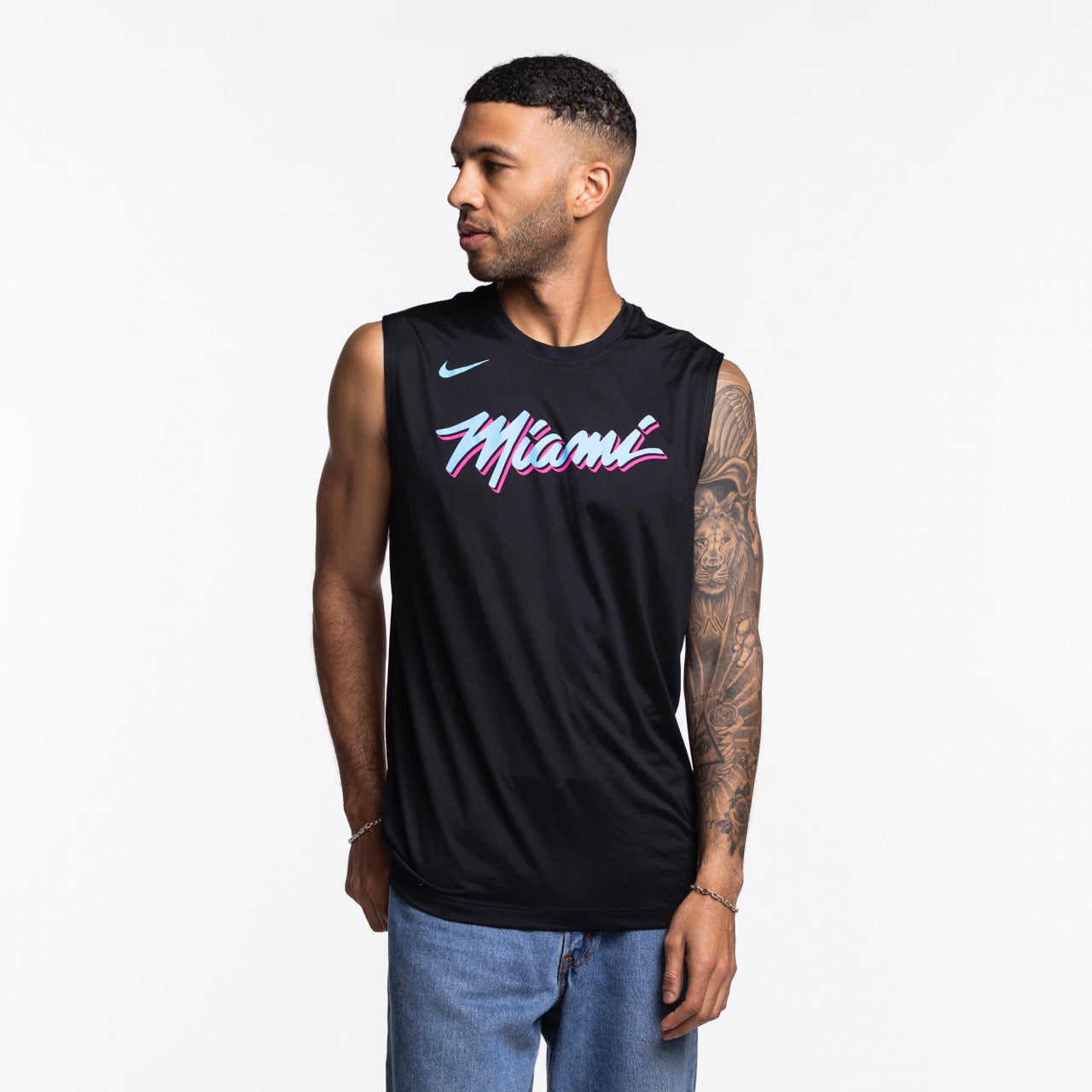 A man with short hair and a tattooed right arm wears a Nike Miami HEAT Original Vice Nights Sleeveless Tee in black and blue jeans. He looks to his left against a plain white background. Wash with like colors.