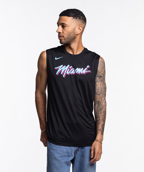 A man with short hair and a tattooed right arm wears a Nike Miami HEAT Original Vice Nights Sleeveless Tee in black and blue jeans. He looks to his left against a plain white background. Wash with like colors.