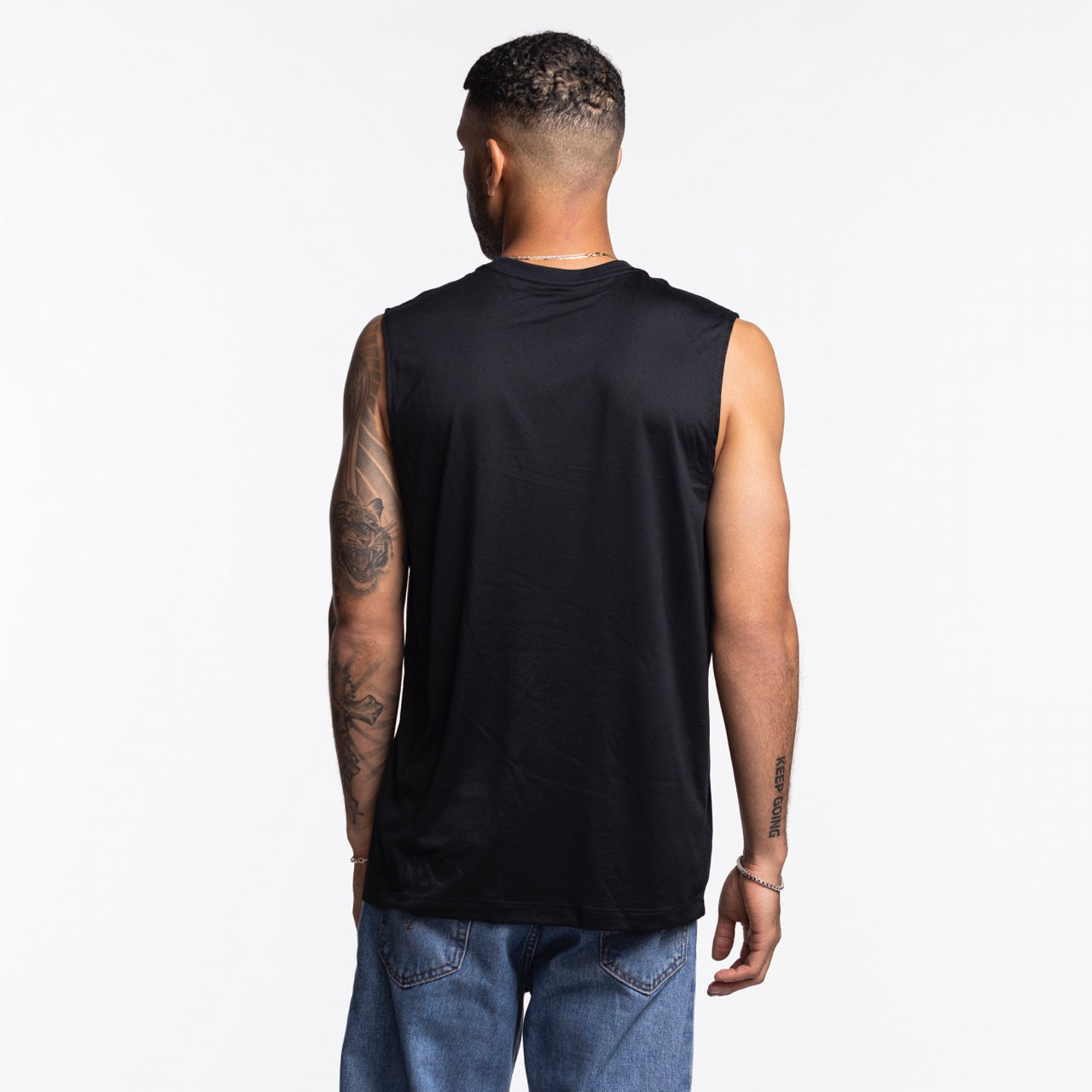 A man with short hair and visible tattoos on his left arm faces away, wearing a Nike Miami HEAT Original Vice Nights Sleeveless Tee in black polyester and blue jeans against a plain white background.