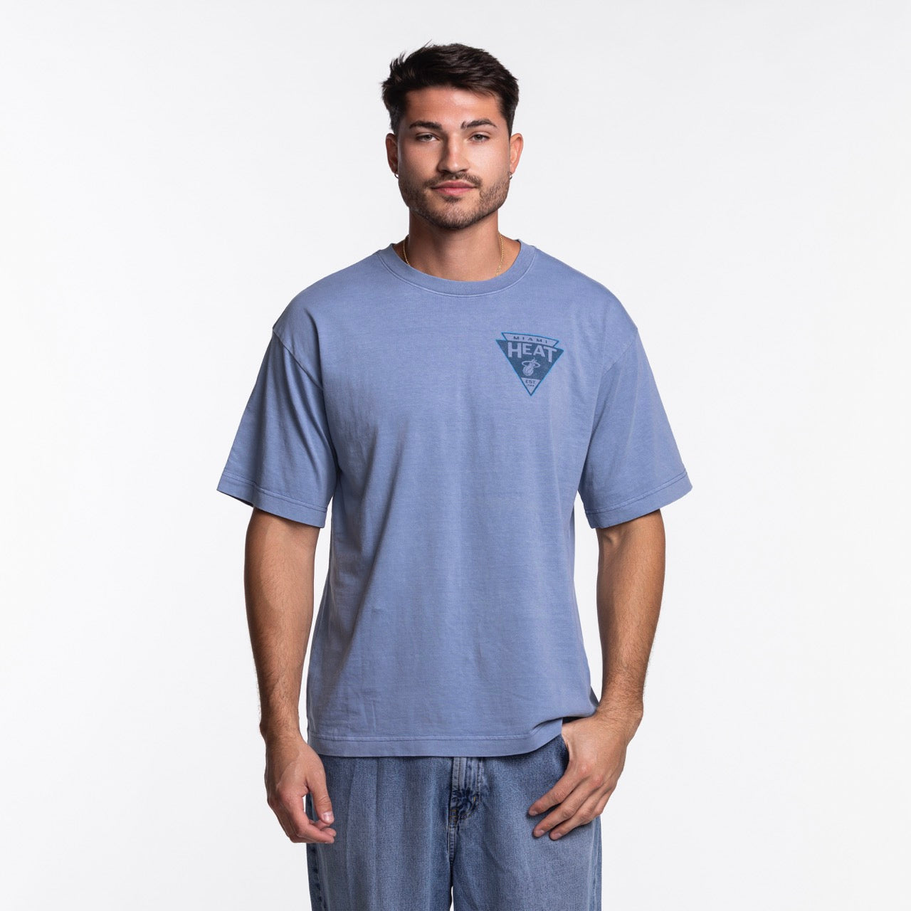 A man with short dark hair and a beard wears the '47 Brand Miami HEAT Original Vice Nights Switchback Tee, a standard fit, light blue 100% cotton shirt with a small HEAT logo on the chest, paired with blue jeans against a white background.