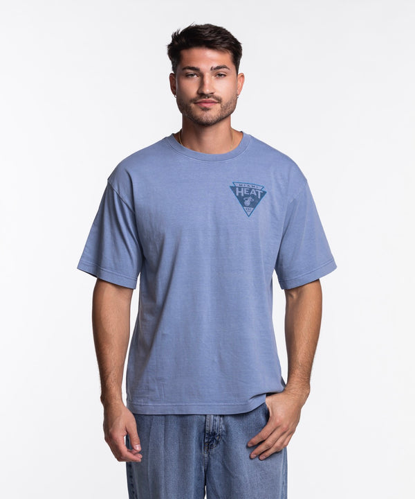 A man with short dark hair and a beard wears the '47 Brand Miami HEAT Original Vice Nights Switchback Tee, a standard fit, light blue 100% cotton shirt with a small HEAT logo on the chest, paired with blue jeans against a white background.