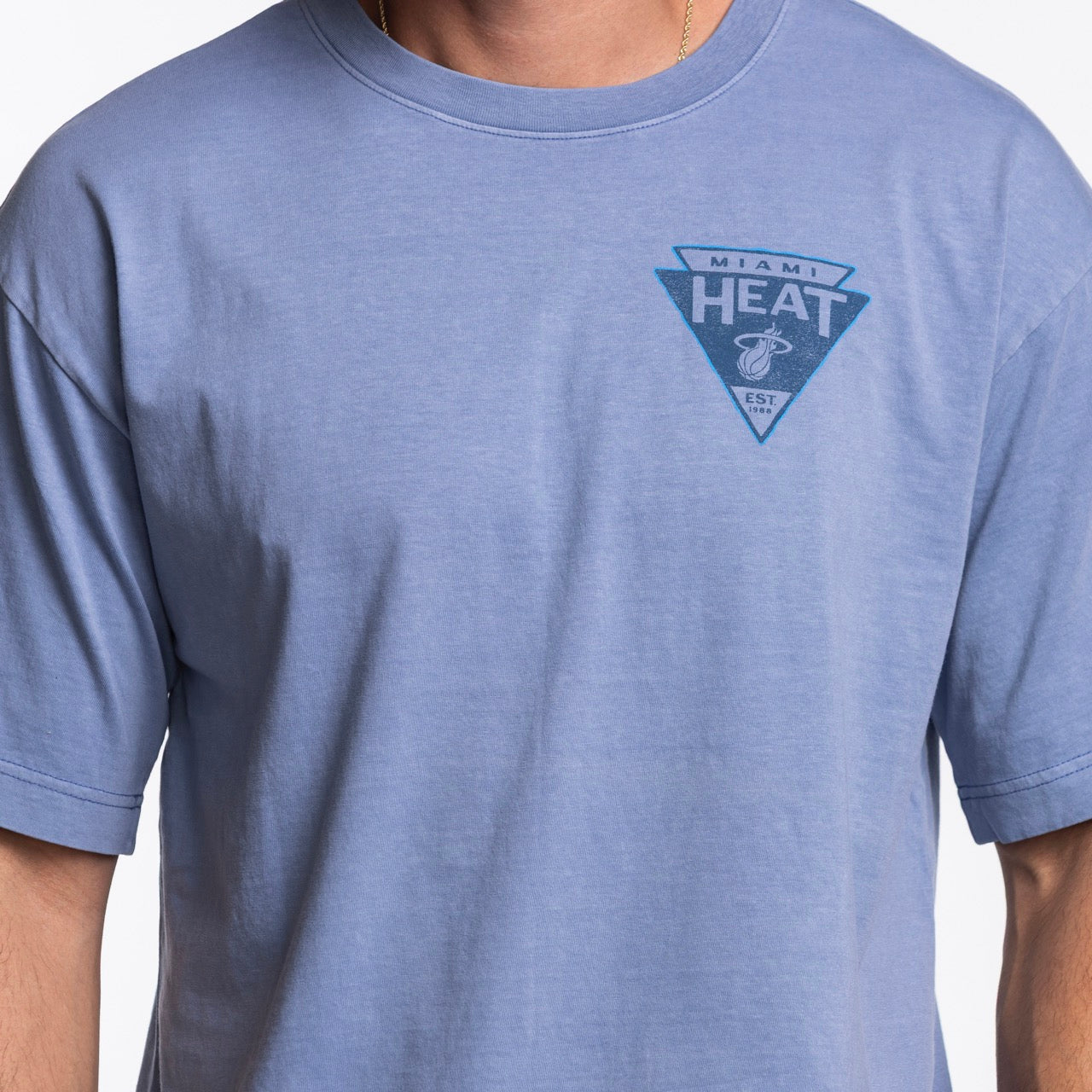 A person wearing the '47 Brand Miami HEAT Original Vice Nights Switchback Tee, a light blue 100% cotton, standard fit shirt featuring a small triangular Miami Heat logo on the left chest. Easy care—machine wash cold.