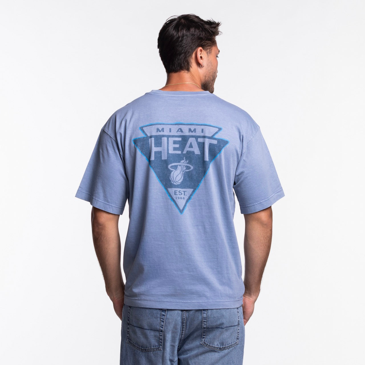 A man in light blue jeans and short dark hair faces away, wearing the '47 Brand Miami HEAT Original Vice Nights Switchback Tee, which features a large team logo and text on the back. The background is plain white.