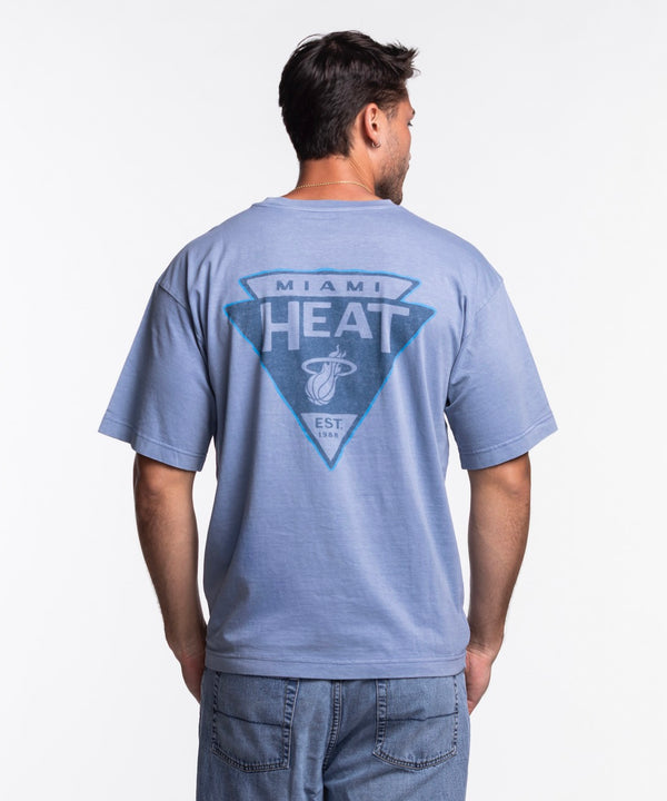 A man with short dark hair and a beard wears the '47 Brand Miami HEAT Original Vice Nights Switchback Tee, a standard fit, light blue 100% cotton shirt with a small HEAT logo on the chest, paired with blue jeans against a white background.