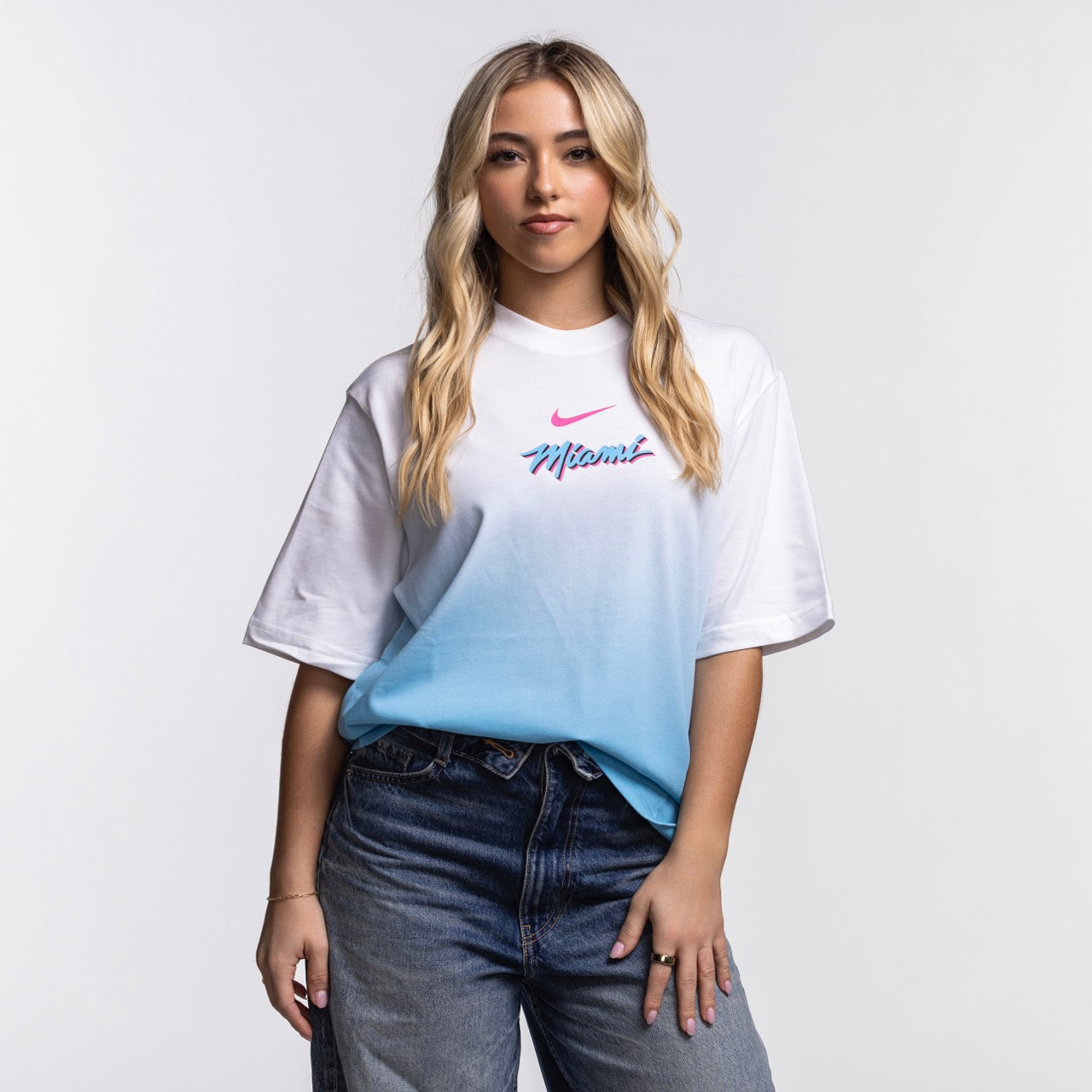 A young woman with long blonde hair stands against a white background, wearing a loose-fit Nike Miami HEAT Original Vice Nights Women's Boxy Tee and blue jeans, with one hand in her pocket and a neutral expression.