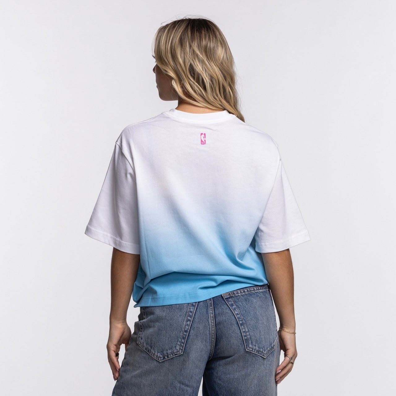 A woman with blonde hair, wearing the Nike Miami HEAT Original Vice Nights Women's Boxy Tee by Nike and blue jeans, stands facing away from the camera against a plain white background.
