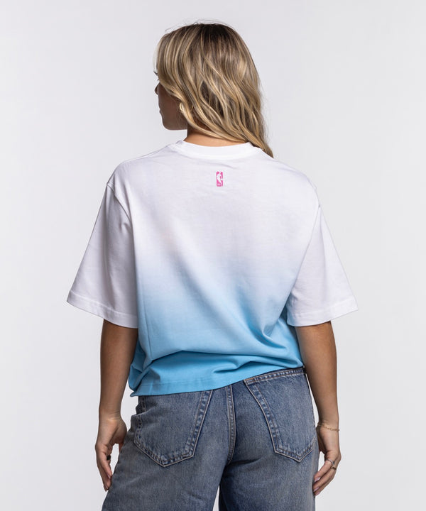 A young woman with long blonde hair stands against a white background, wearing a loose-fit Nike Miami HEAT Original Vice Nights Women's Boxy Tee and blue jeans, with one hand in her pocket and a neutral expression.