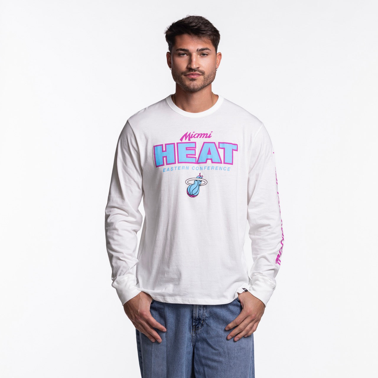 A man faces forward in blue jeans and a '47 Brand Miami HEAT Original Vice Nights White Long Sleeve Tee featuring pink and blue lettering with a basketball logo on the front. The background is plain white.
