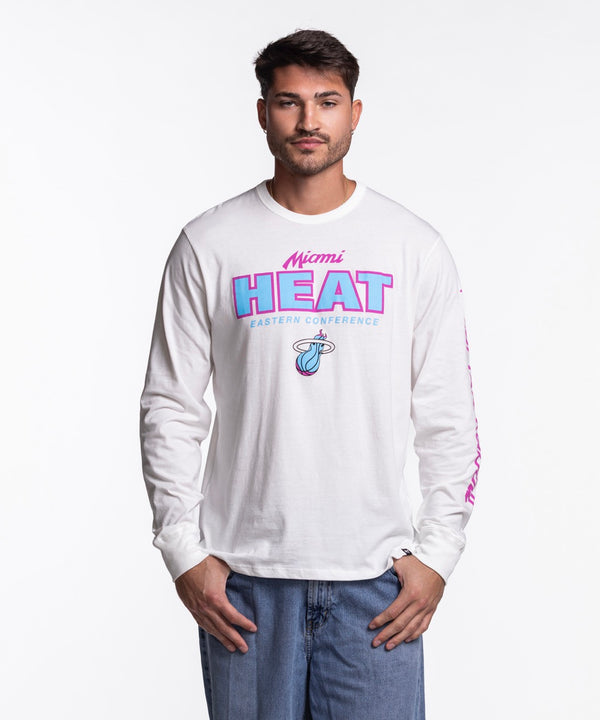A man faces forward in blue jeans and a '47 Brand Miami HEAT Original Vice Nights White Long Sleeve Tee featuring pink and blue lettering with a basketball logo on the front. The background is plain white.