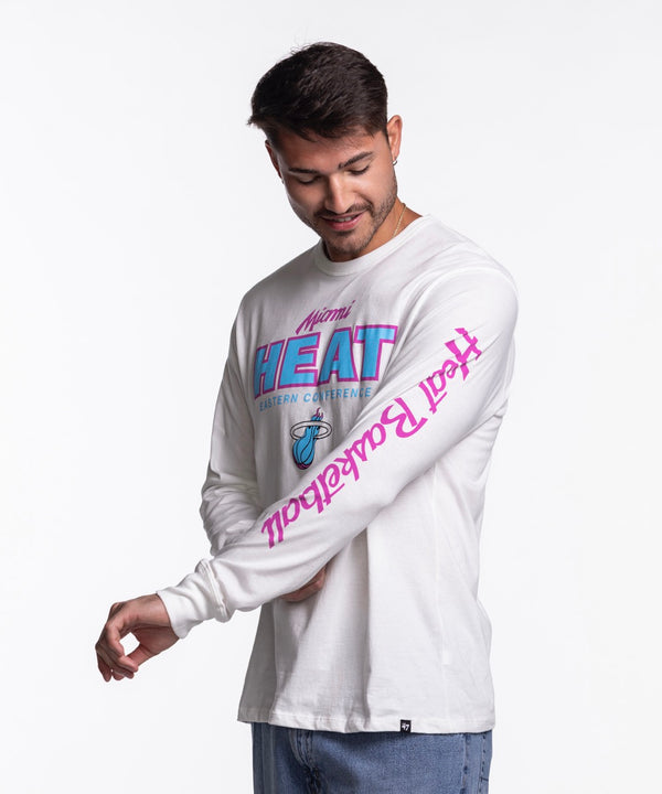 A man faces forward in blue jeans and a '47 Brand Miami HEAT Original Vice Nights White Long Sleeve Tee featuring pink and blue lettering with a basketball logo on the front. The background is plain white.