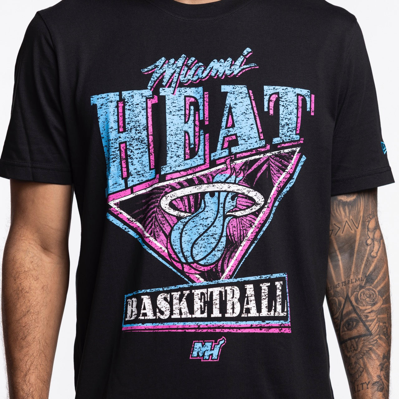 A person with a tattooed arm wears the New Era Miami HEAT Original Vice Nights Graphic Tee, a soft cotton t-shirt featuring retro pink, teal, and white graphics plus the team name and logo.