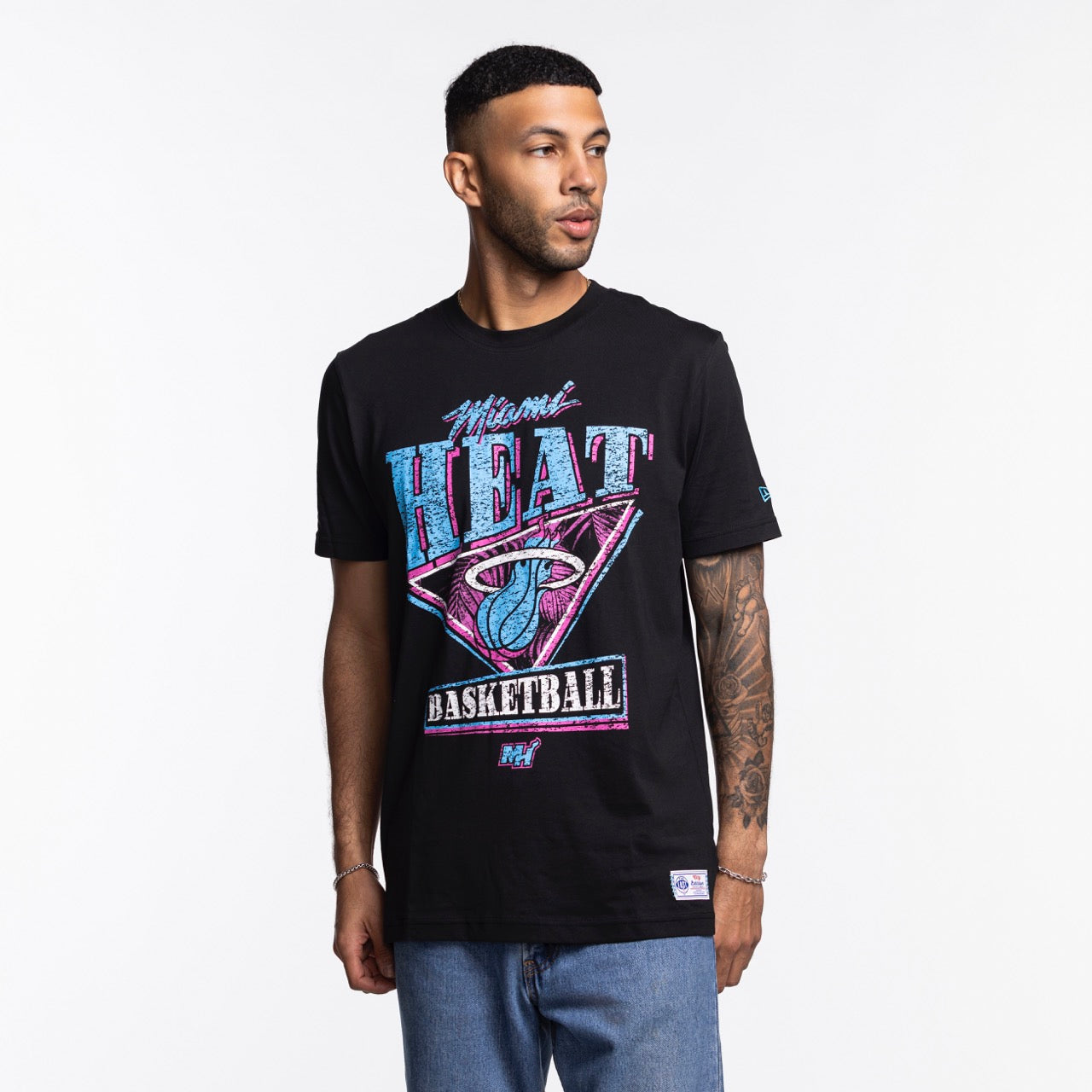 A man in a New Era Miami HEAT Original Vice Nights Graphic Tee with colorful retro graphics stands against a white background. The soft cotton, standard fit shirt is machine washable. He has short hair, tattoos on one arm, and wears blue jeans.