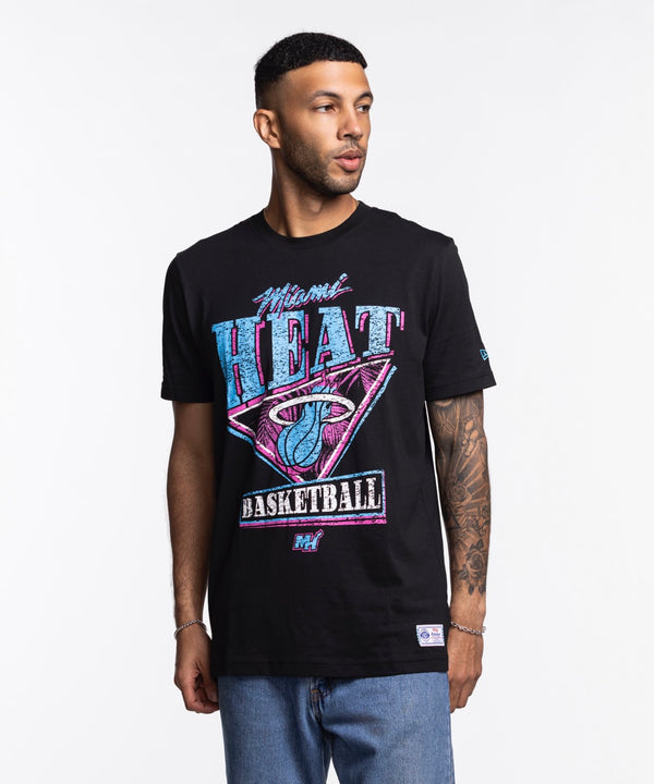A man in a New Era Miami HEAT Original Vice Nights Graphic Tee with colorful retro graphics stands against a white background. The soft cotton, standard fit shirt is machine washable. He has short hair, tattoos on one arm, and wears blue jeans.