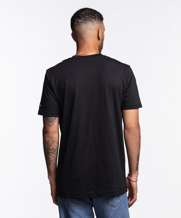 A man in a New Era Miami HEAT Original Vice Nights Graphic Tee with colorful retro graphics stands against a white background. The soft cotton, standard fit shirt is machine washable. He has short hair, tattoos on one arm, and wears blue jeans.