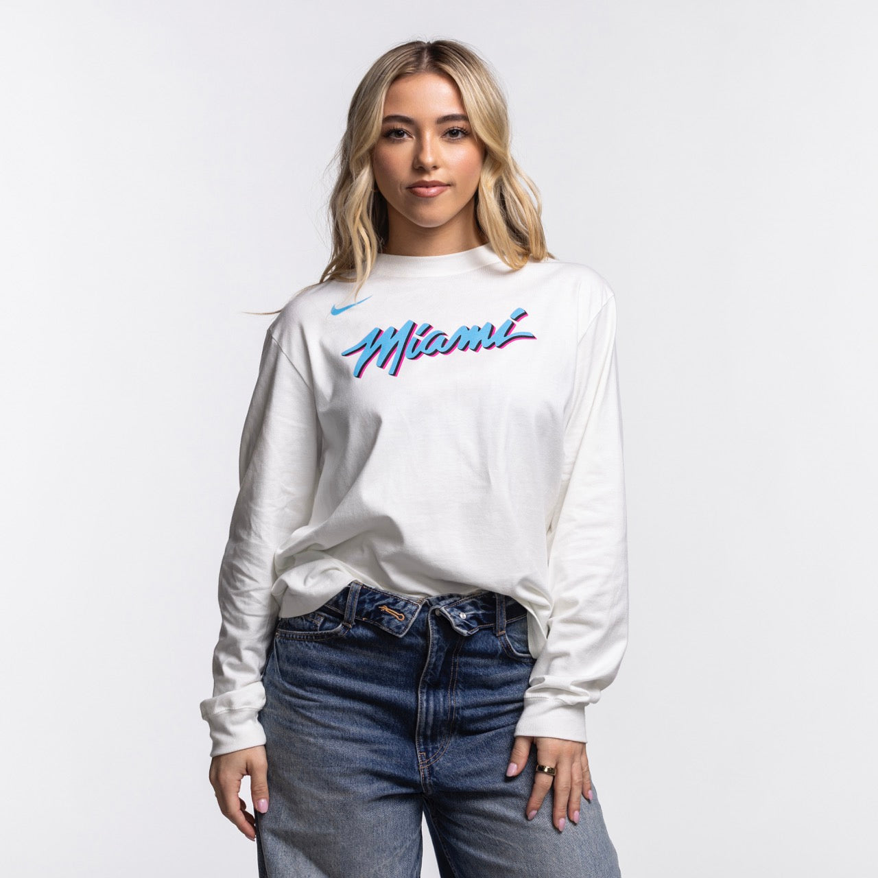A woman with long blonde hair wears a Nike Miami HEAT Original Vice Nights Women's Long Sleeve Tee and blue jeans, standing against a plain white background and facing the camera with a slight smile.