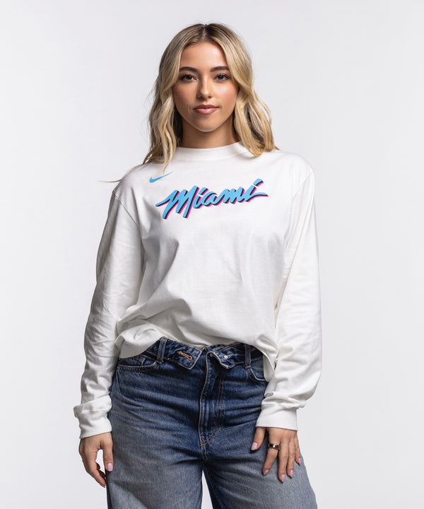 A woman with long blonde hair wears a Nike Miami HEAT Original Vice Nights Women's Long Sleeve Tee and blue jeans, standing against a plain white background and facing the camera with a slight smile.