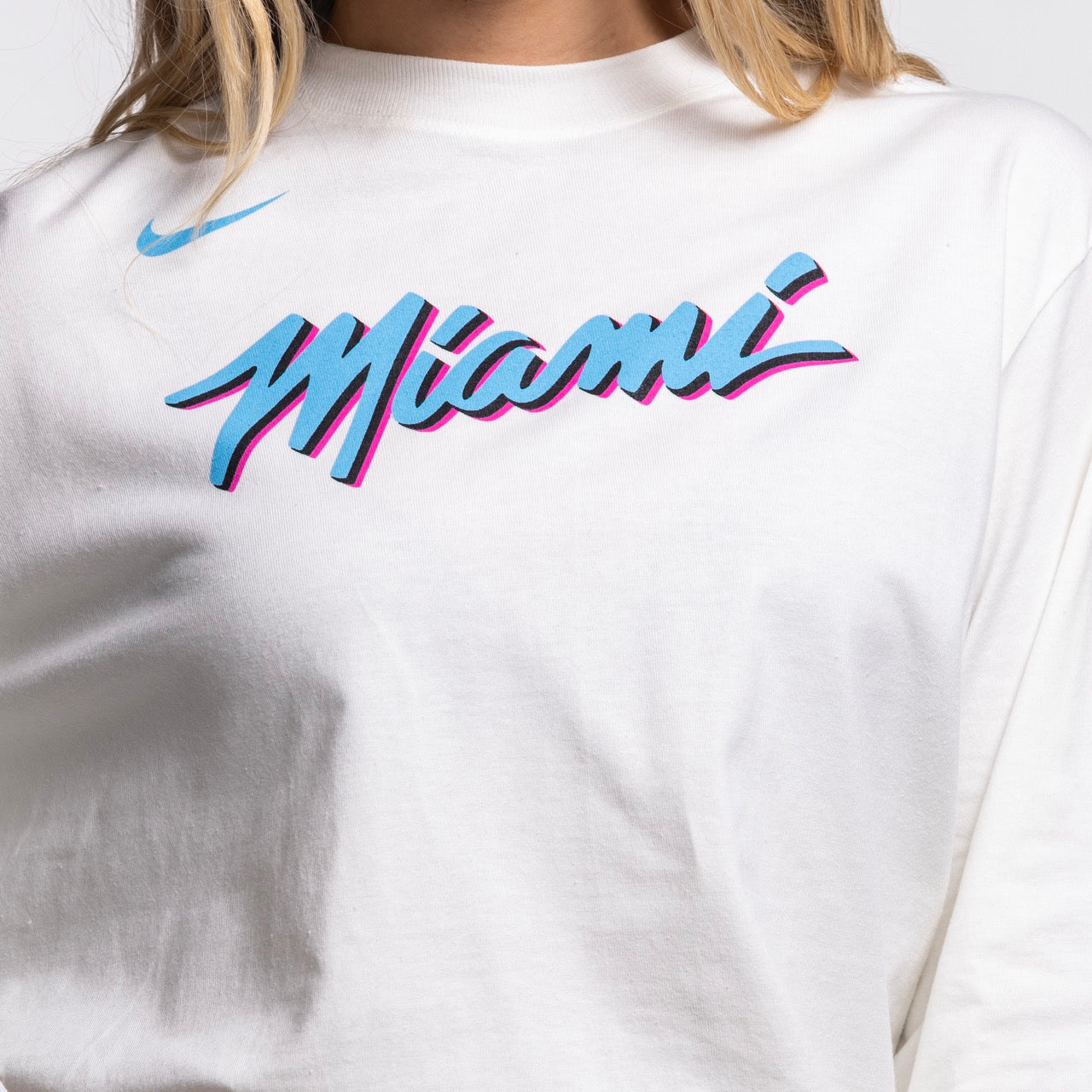 A woman wears the Nike Miami HEAT Original Vice Nights Women's Long Sleeve Tee, a loose-fitting white cotton shirt featuring 