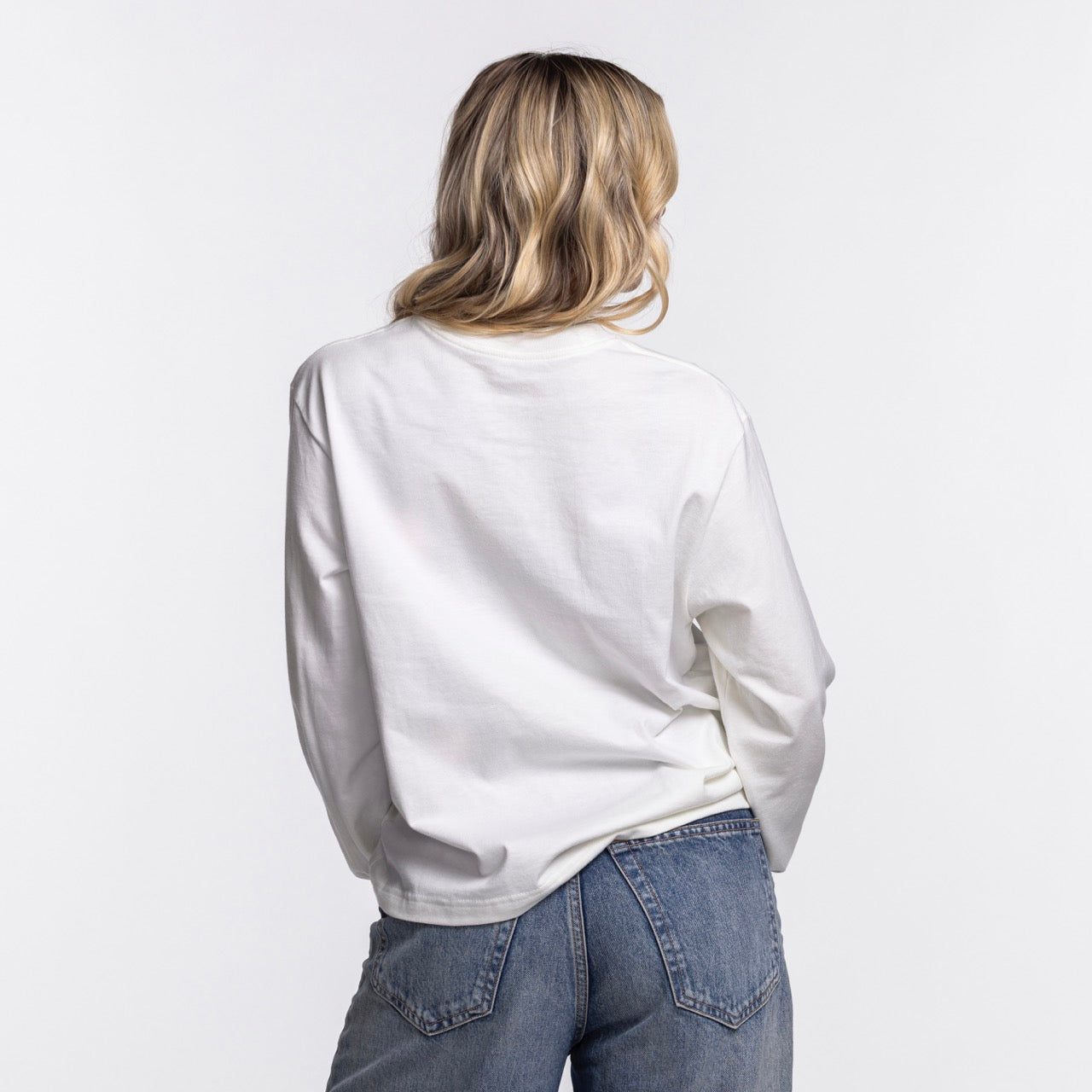 A woman with long, wavy blonde hair is seen from behind wearing the Nike Miami HEAT Original Vice Nights Women's Long Sleeve Tee and blue jeans against a plain white background.