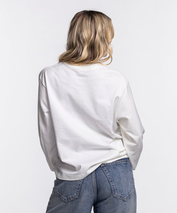 A woman with long blonde hair wears a Nike Miami HEAT Original Vice Nights Women's Long Sleeve Tee and blue jeans, standing against a plain white background and facing the camera with a slight smile.