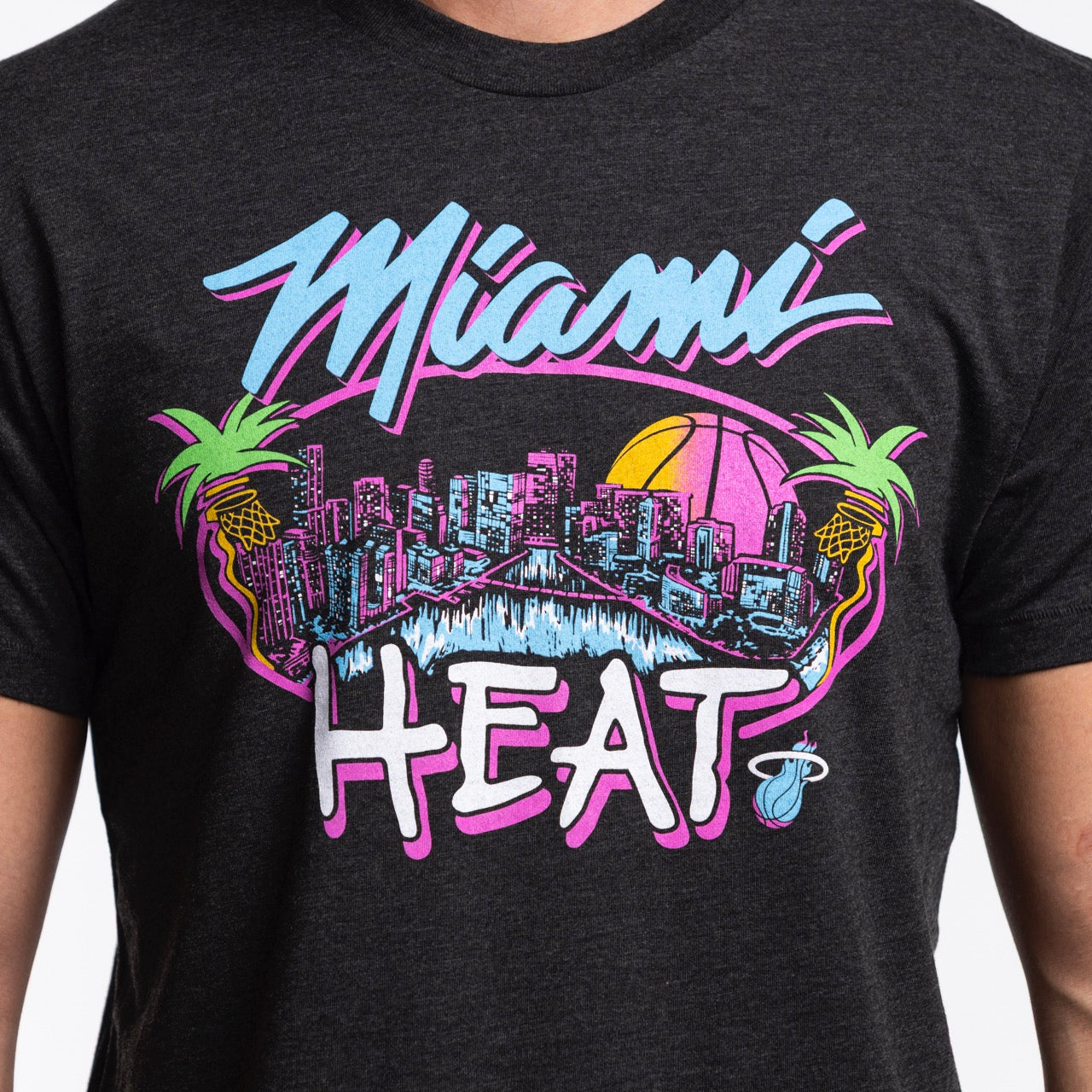 Someone wearing the Homage Miami HEAT Original Vice Nights City Tee by Homage, a black shirt featuring vibrant neon city skyline, palm trees, and a sunset basketball with colorful 