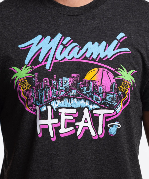 A man wears the Homage Miami HEAT Original Vice Nights City Tee by Homage, a black graphic t-shirt, standing against a plain white background and looking slightly to the side with a relaxed expression.