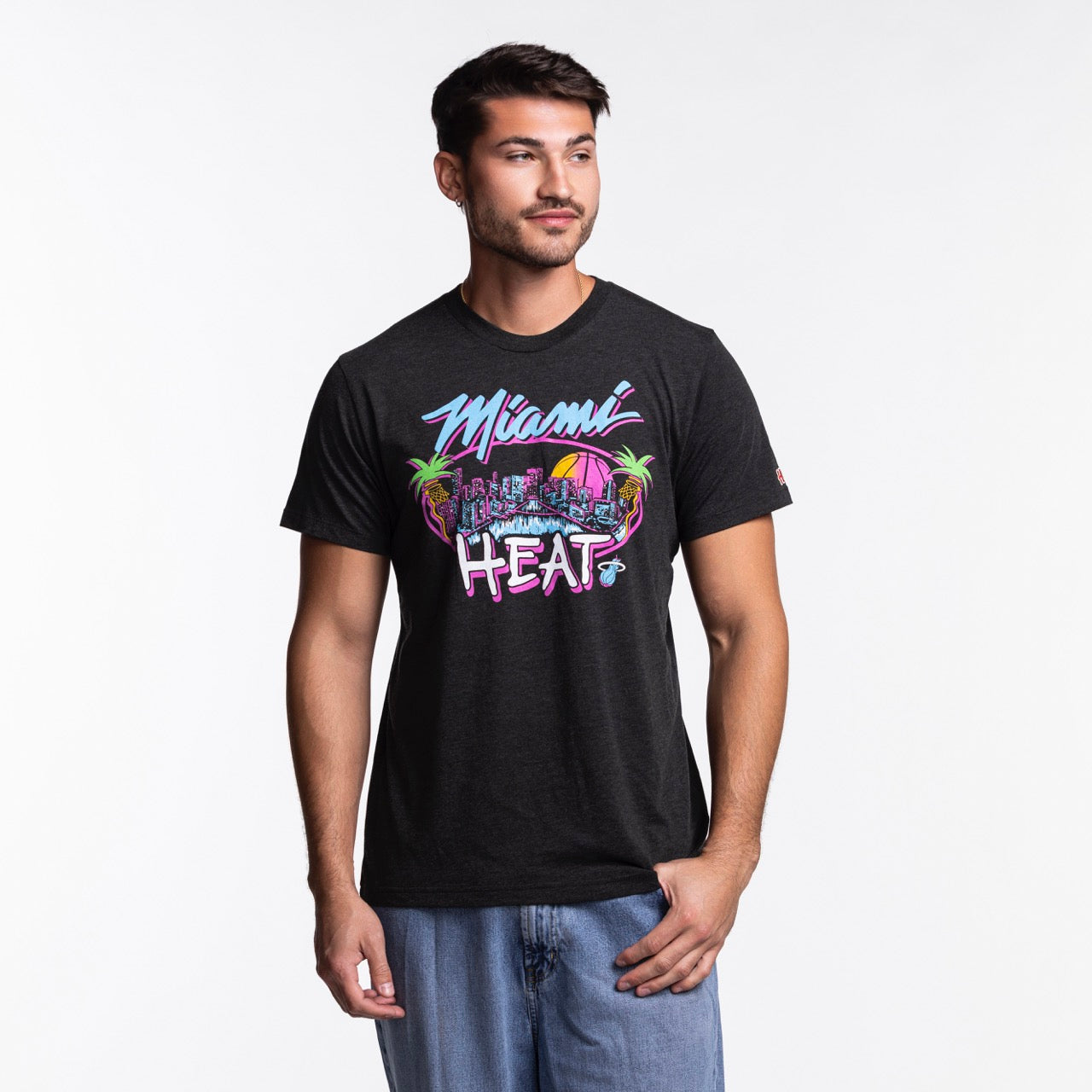 A man wears the Homage Miami HEAT Original Vice Nights City Tee by Homage, a black graphic t-shirt, standing against a plain white background and looking slightly to the side with a relaxed expression.