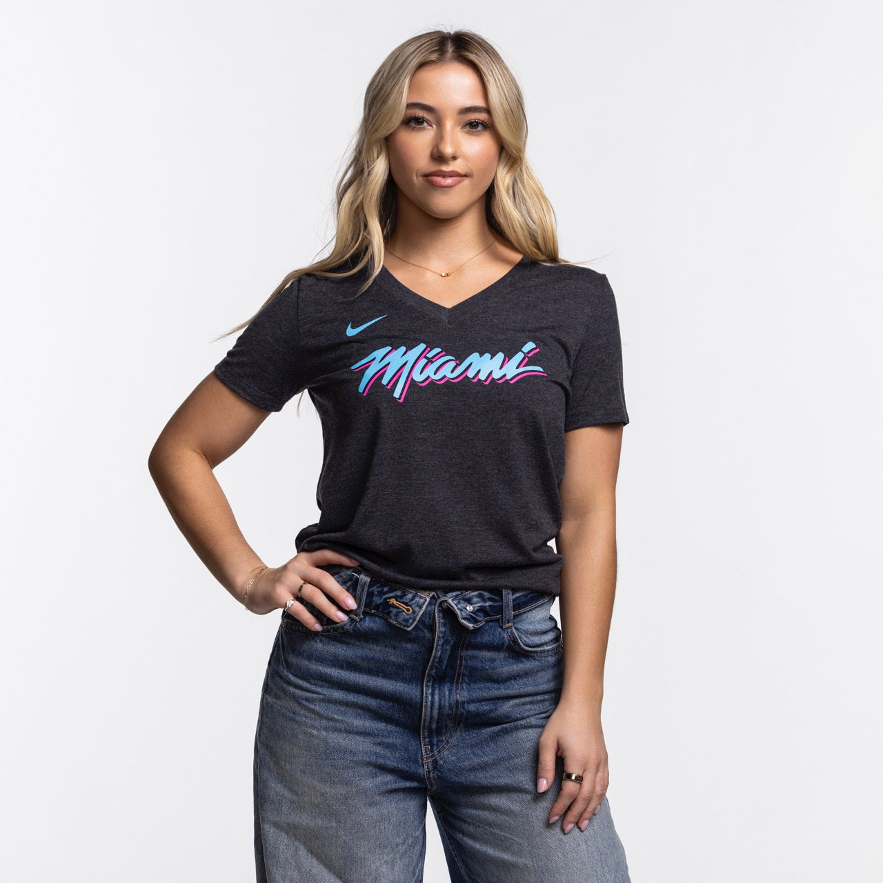 A woman with long blonde hair wears the Nike Miami HEAT Original Vice Nights Women's V-Neck Tee in dark gray with colorful 
