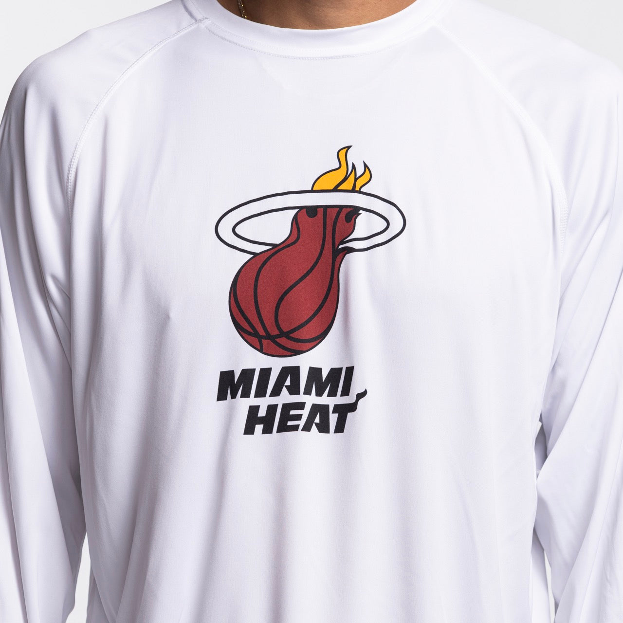 A person wears the Columbia Outdoor Miami HEAT Logo Terminal Long Sleeve Tee, a white polyester shirt with the team's flaming basketball and MIAMI HEAT text. It's standard fit and machine washable.