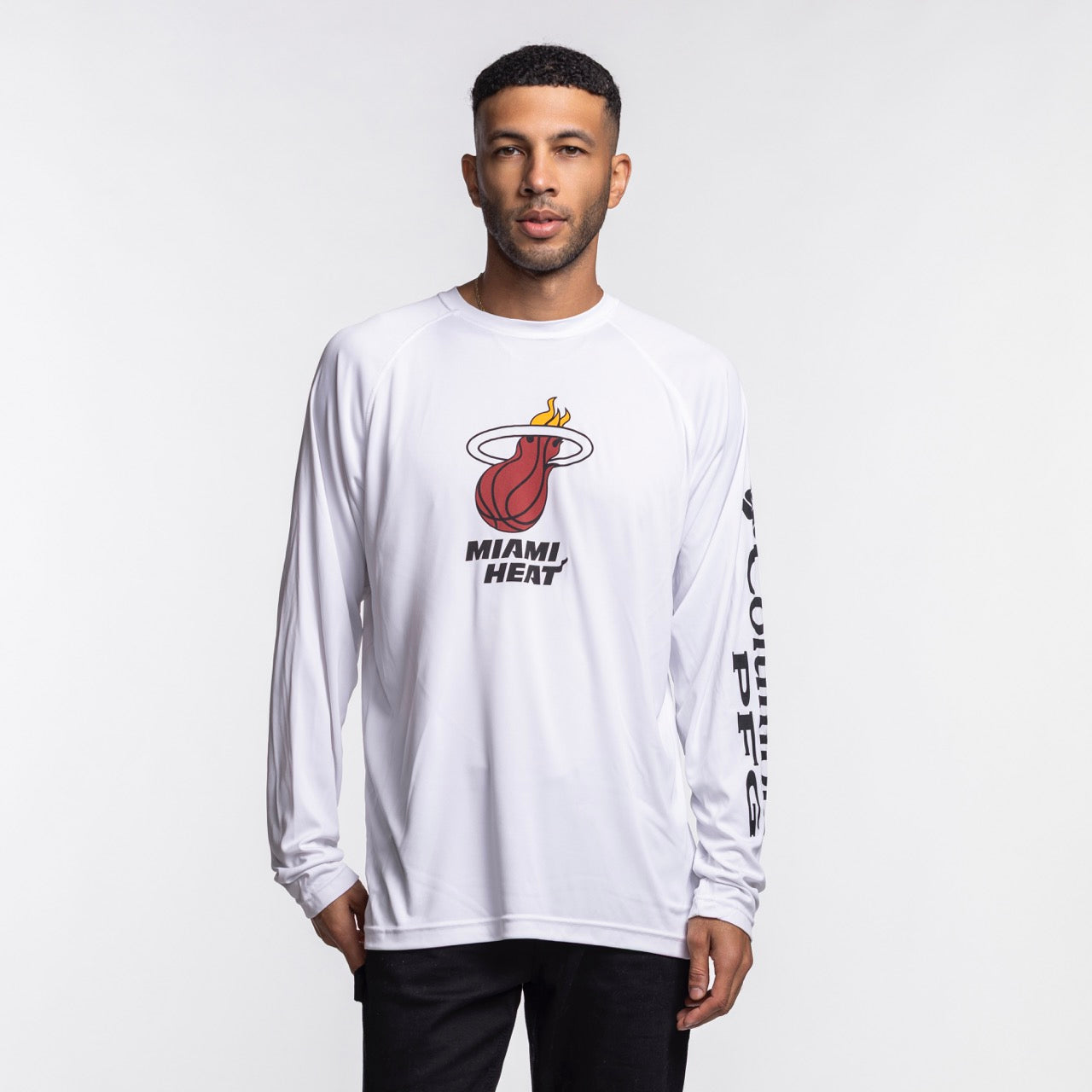 A man wearing a Columbia Miami HEAT Logo Terminal Long Sleeve Tee by Columbia Outdoor and black pants stands against a plain white background.