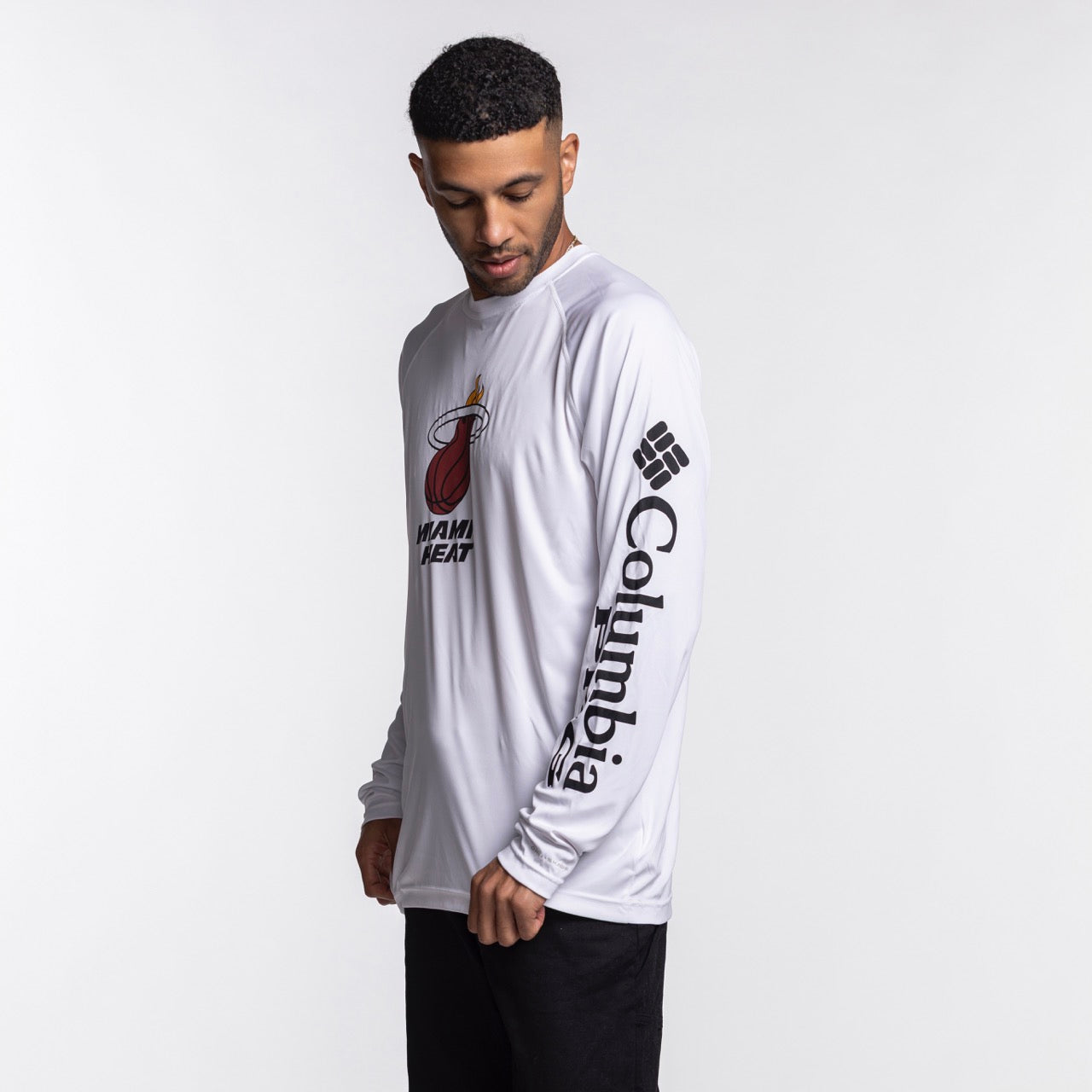 A man stands against a plain white background wearing the Columbia Miami HEAT Logo Terminal Long Sleeve Tee by Columbia Outdoor, featuring a white polyester fabric, standard fit, Miami Heat logo on the chest, and Columbia branding on the sleeve.