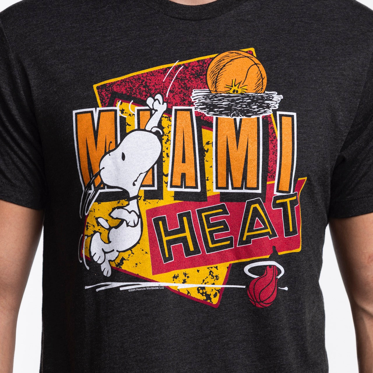 The Homage Miami HEAT Snoopy Tee by Homage is a soft black shirt featuring Snoopy from Peanuts dunking a basketball, with bold MIAMI HEAT text and the team logo below in yellow, orange, red, and white.