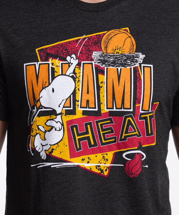 A man wears the Homage Miami HEAT Snoopy Tee by Homage—a black graphic t-shirt—paired with blue jeans, standing against a white background, looking right with a slight smile and his hands by his sides.
