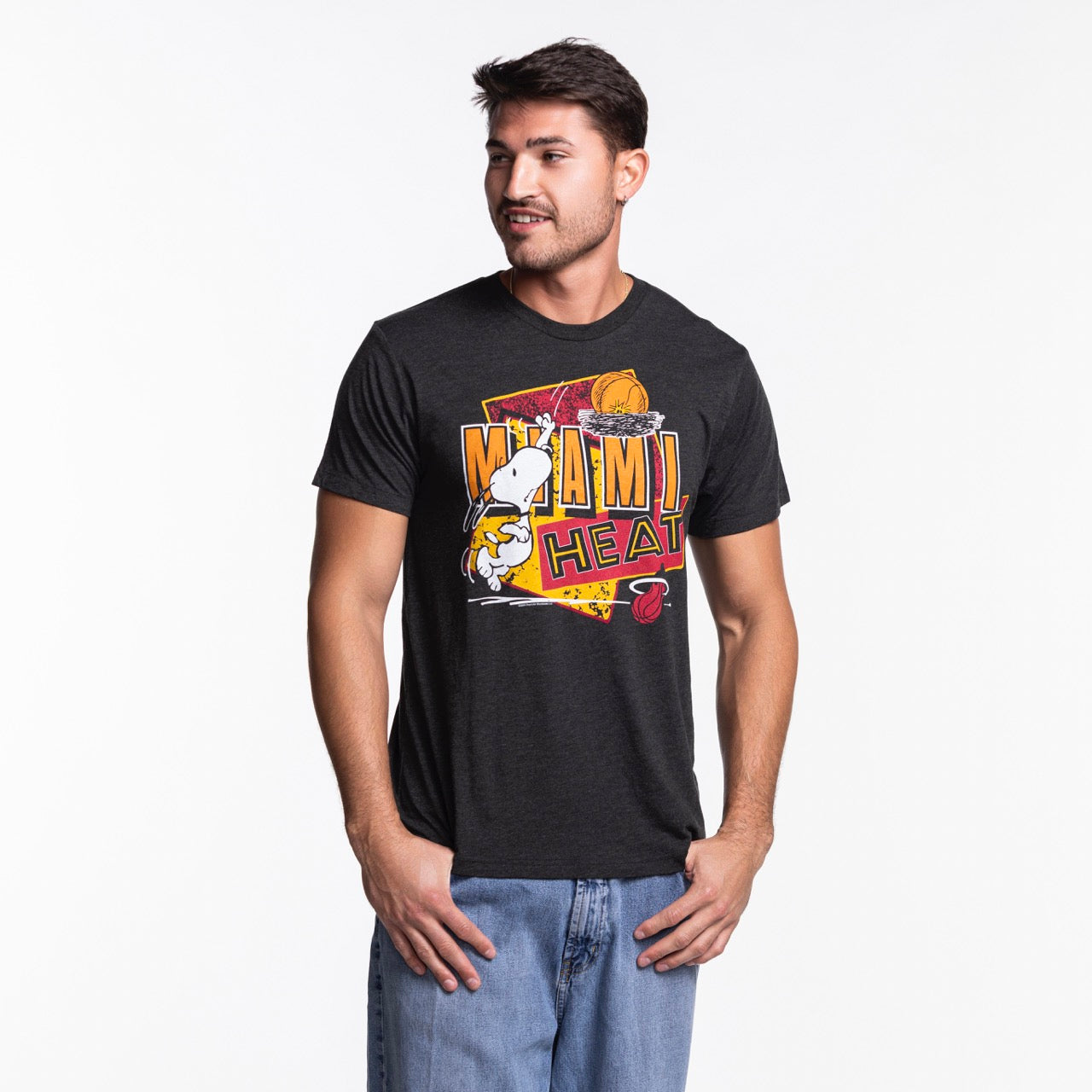 A man wears the Homage Miami HEAT Snoopy Tee by Homage—a black graphic t-shirt—paired with blue jeans, standing against a white background, looking right with a slight smile and his hands by his sides.