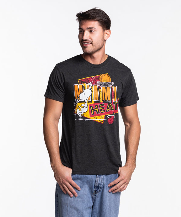 A man wears the Homage Miami HEAT Snoopy Tee by Homage—a black graphic t-shirt—paired with blue jeans, standing against a white background, looking right with a slight smile and his hands by his sides.