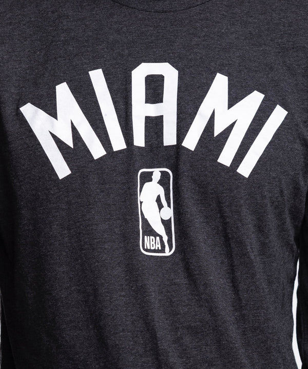 A man wears the Sportiqe Miami HEAT NBA Long Sleeve Tee—a black shirt by Sportiqe featuring MIAMI and an NBA logo. Made from a polyester-cotton-rayon blend, he stands against a white background, looking left with one hand in his pocket.